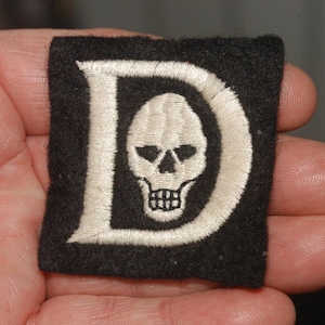 May include: A black fabric patch with a white embroidered letter "D" and a skull inside the letter. The skull is white with black eyes and a black mouth.