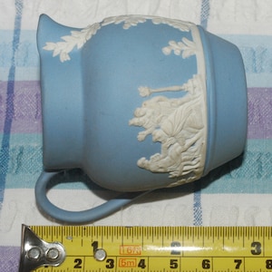 May include: A light blue ceramic pitcher with white relief detailing of a scene with figures. The pitcher has a handle and measures 4 inches in length.