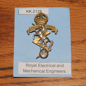 May include: A silver and gold metal emblem with a crown, horse, and lightning bolt design. The emblem is set on a blue background. The text "Royal Electrical and Mechanical Engineers" is below the emblem. The text "KK.2128" is above the emblem.