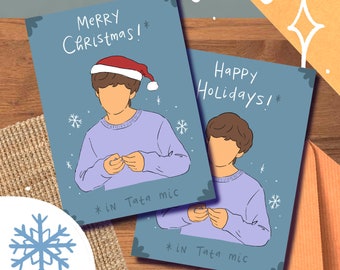 May include: Two Christmas cards featuring a cartoon illustration of a person wearing a purple sweater and a Santa hat. The text on the cards reads "Merry Christmas!" and "Happy Holidays!" with the phrase "* in Tata mic" at the bottom of each card.