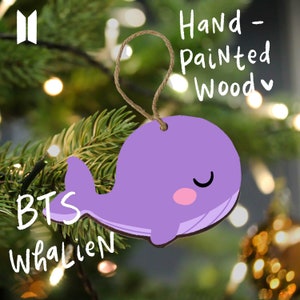 Könnte beinhalten: Lilafarbener, handbemalter Holz-Anhänger in Form eines Wals mit dem Text "BTS Whalien" auf einem Weihnachtsbaum. Der Anhänger ist mit "Handbemaltes Holz" beschriftet.