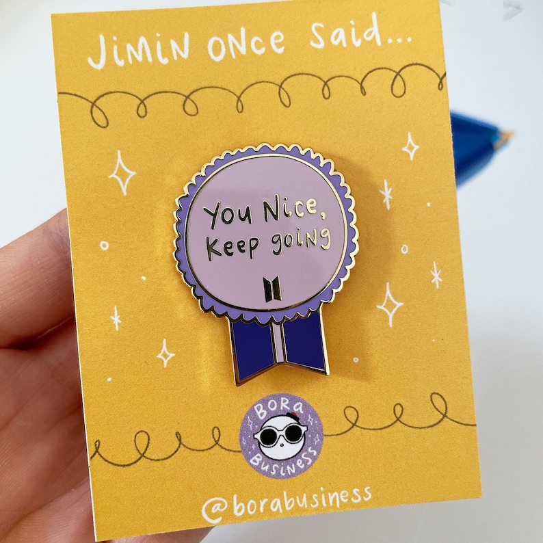 May include: A gold-coloured enamel pin with a purple background and the text "You Nice, Keep going" in gold lettering. The pin features a stylised BTS logo and a blue ribbon. The pin is on a yellow background with the text "Jimin Once Said..." and a small cartoon bear with sunglasses and the text "Bora Business" and "@borabusiness".