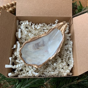 May include: A natural oyster shell with a gold-painted rim, presented in a brown cardboard box filled with white paper filler. The shell's interior has a pearlescent white and tan color. The box is open, revealing the shell and filler.