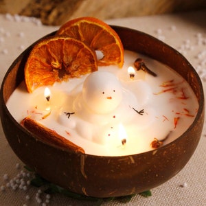 May include: A white snowman candle with orange slices and cinnamon sticks in a brown coconut shell. The candle is lit and the wax is melted.