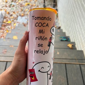 May include: A pink and white tumbler with a cartoon character holding a can of Coca-Cola. The text on the tumbler reads "Tomando Coca Mi riñón se relaja".