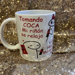 May include: White ceramic mug with a red and black cartoon illustration and the text "Tomando Coca Mi riñón se relaja" and "Y Más Menos A".