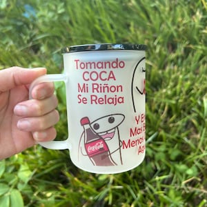 May include: White ceramic mug with a black rim and handle. The mug has a cartoon illustration of a kidney drinking a Coca-Cola bottle. The text on the mug reads "Tomando Coca Mi Riñón Se Relaja" and "Y En Mas B Menos A".