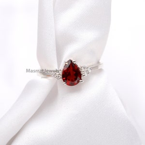 Może przedstawiać: Pierścionek z czerwonym kamieniem szlachetnym w kształcie gruszki osadzonym w srebrze, z mniejszymi, przezroczystymi kamieniami na obrączce.
