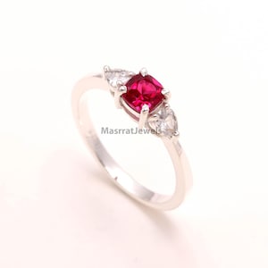Può includere: Un anello d'argento con una gemma rossa quadrata e vibrante al centro, affiancata da due gemme bianche a forma di pera più piccole. L'anello ha una fascia delicata ed è posto su uno sfondo bianco. Il testo "MasrratJewels" è visibile sull'anello.