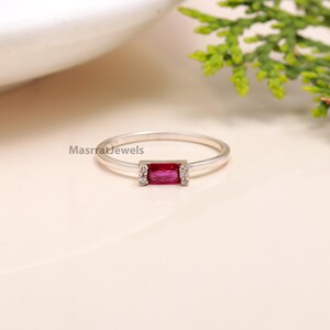 May include: A silver ring with a rectangular red gemstone and small clear gemstones on either side. MasrratJewels is printed on the band.