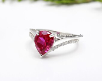 Peut inclure: Une bague en argent avec une pierre précieuse en rubis de forme poire et une rangée de petits diamants sur la bande. La bague est posée sur un fond blanc.