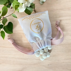 May include: A white drawstring bag with a pink ribbon tie. The bag is filled with small, light green ceramic beads. The bag has a gold logo that says "Essentials" with a leaf design. The text on the bag reads "Jasmine & Green Floral Scented Ceramic Beads".