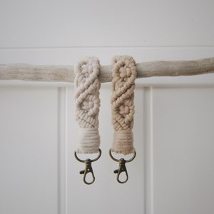 May include: Two beige macrame keychains with a gold metal key ring. The keychains are hanging from a branch.