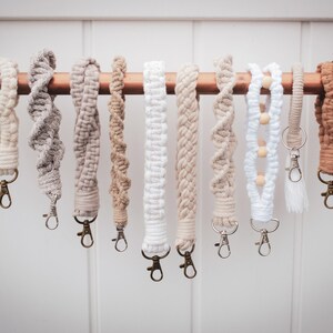 May include: A collection of 10 handmade macrame keychains in various shades of beige and white. The keychains are hanging from a copper pipe and feature different patterns and textures.