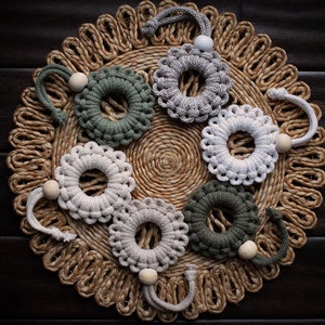 May include: Six crocheted macrame wall hangings in shades of green and white with wooden beads. The hangings are arranged in a circle on a woven straw mat.