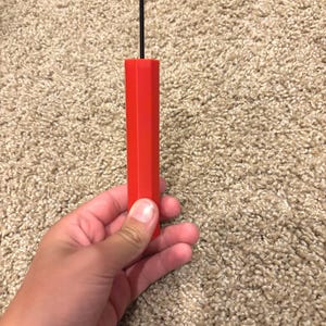 Puede incluir: Un objeto rectangular rojo con una línea negra fina y vertical que se extiende desde la parte superior. El objeto se sostiene en una mano. El fondo es una superficie texturizada de color marrón claro.