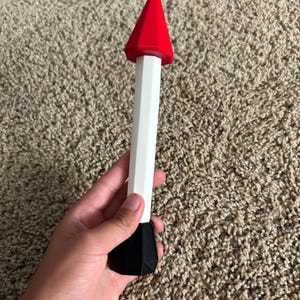 Puede incluir: Objeto decorativo blanco, negro y rojo con diseño geométrico. Presenta una parte superior cónica roja, un cuerpo blanco y una base negra. El objeto se sostiene en una mano.