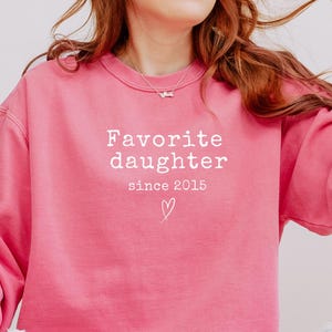 Pode incluir: Um moletom rosa vibrante com o texto branco "Favorite daughter since 2015" e um s&iacute;mbolo de cora&ccedil;&atilde;o. O moletom &eacute; de cor s&oacute;lida e tem um estilo casual.