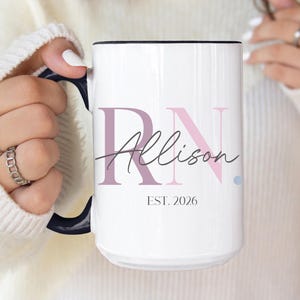 May include: A white ceramic mug with a dark blue handle and rim. The front features the large letters RN in muted pink and purple, with the name Allison written in black script across the center and the text EST. 2026 printed in black below.