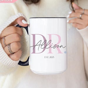 May include: White ceramic mug with a black interior and handle. The mug features the text "DR Allison" in a stylish font, with the year "EST. 2025" below. The mug is held by a person wearing a white sweater.