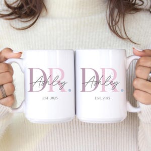 May include: Two white ceramic mugs with the text "DR. Ashley EST. 2025" printed in a stylish font. The "DR" is in a mauve color, and "Ashley" is in a script font. A small blue dot is next to the name.