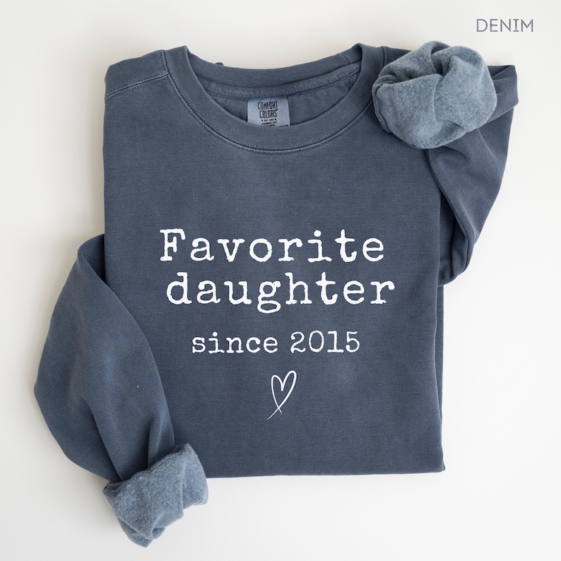 Pode incluir: Um moletom azul denim com as palavras "Favorite daughter since 2015" impressas em branco. Um pequeno cora&ccedil;&atilde;o &eacute; desenhado abaixo do texto. O moletom tem uma manga dobrada e uma etiqueta Comfort Colors.