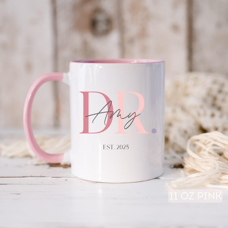 May include: White ceramic mug with a pink interior and handle. The mug features the text "Dr. Amy" in pink and gray, with "EST. 2025" below. The bottom right corner of the mug reads "11 OZ PINK".
