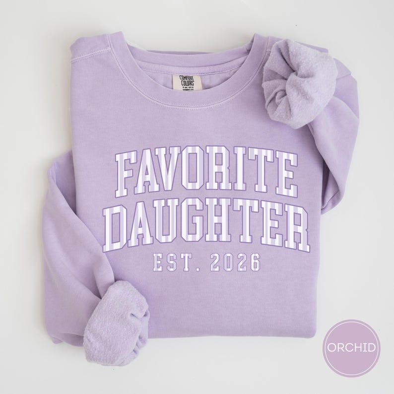 May include: A light purple crewneck sweatshirt featuring the text FAVORITE DAUGHTER EST. 2026 in white block lettering with vertical stripes. The garment has long sleeves with gathered cuffs and a Comfort Colors brand label at the inner neckline.
