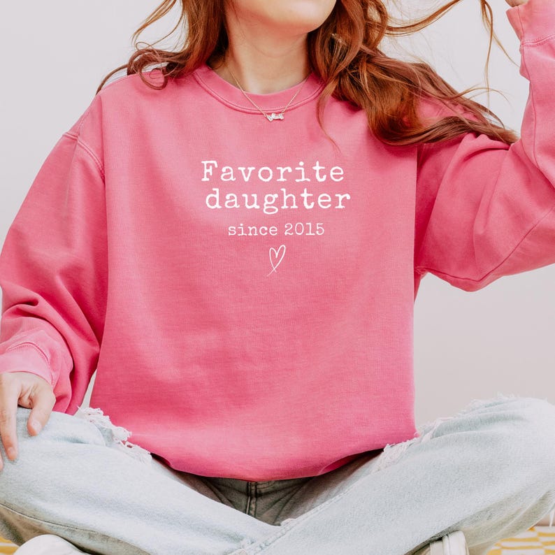 Pode incluir: Um moletom rosa vibrante com o texto branco "Favorite daughter since 2015" e um s&iacute;mbolo de cora&ccedil;&atilde;o. O moletom &eacute; de cor s&oacute;lida e tem um estilo casual.