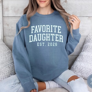 May include: A blue crewneck sweatshirt featuring the white text FAVORITE DAUGHTER EST. 2026 printed in a collegiate font on the chest. The garment has long sleeves, a ribbed neckline, and a relaxed fit, showcasing a solid blue color throughout.