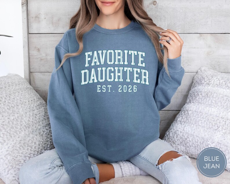 May include: A blue crewneck sweatshirt featuring the white text FAVORITE DAUGHTER EST. 2026 printed in a collegiate font on the chest. The garment has long sleeves, a ribbed neckline, and a relaxed fit, showcasing a solid blue color throughout.