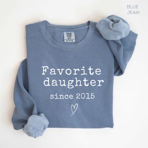 Pode incluir: Camisola azul jeans com as palavras "Favorite daughter since 2015" em branco e um pequeno cora&ccedil;&atilde;o. A camisola tem gola redonda e mangas compridas. A etiqueta da marca &eacute; vis&iacute;vel no topo.