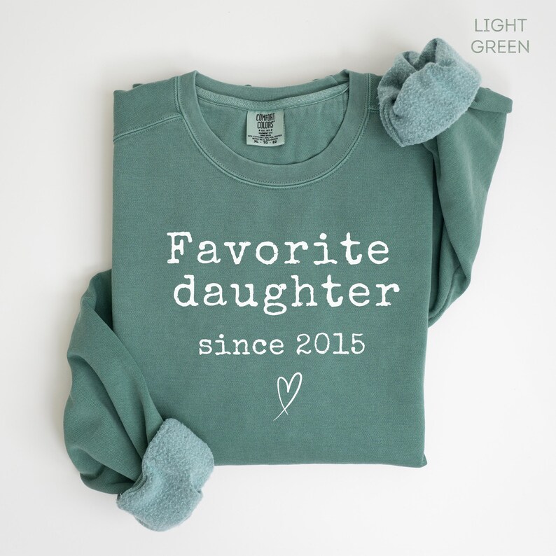 Pode incluir: Camisola verde claro com as palavras "Favorite daughter since 2015" em letras brancas. Um pequeno cora&ccedil;&atilde;o branco est&aacute; abaixo do texto. As mangas est&atilde;o enroladas, mostrando o interior verde claro. Uma pe&ccedil;a confort&aacute;vel.