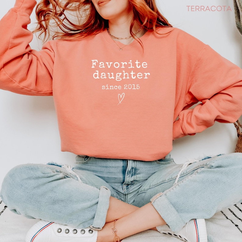 Pode incluir: Um moletom cor de terracota com o texto branco "Favorite daughter since 2015". O moletom tem gola redonda e mangas compridas. Um pequeno s&iacute;mbolo de cora&ccedil;&atilde;o acompanha o texto.