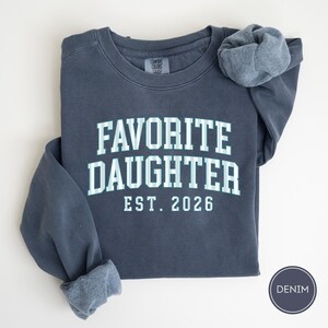 May include: A dark blue crewneck sweatshirt featuring the text FAVORITE DAUGHTER EST. 2026 in a light blue and white striped font. The garment has a Comfort Colors brand label at the inner neckline and a circular badge labeled DENIM.