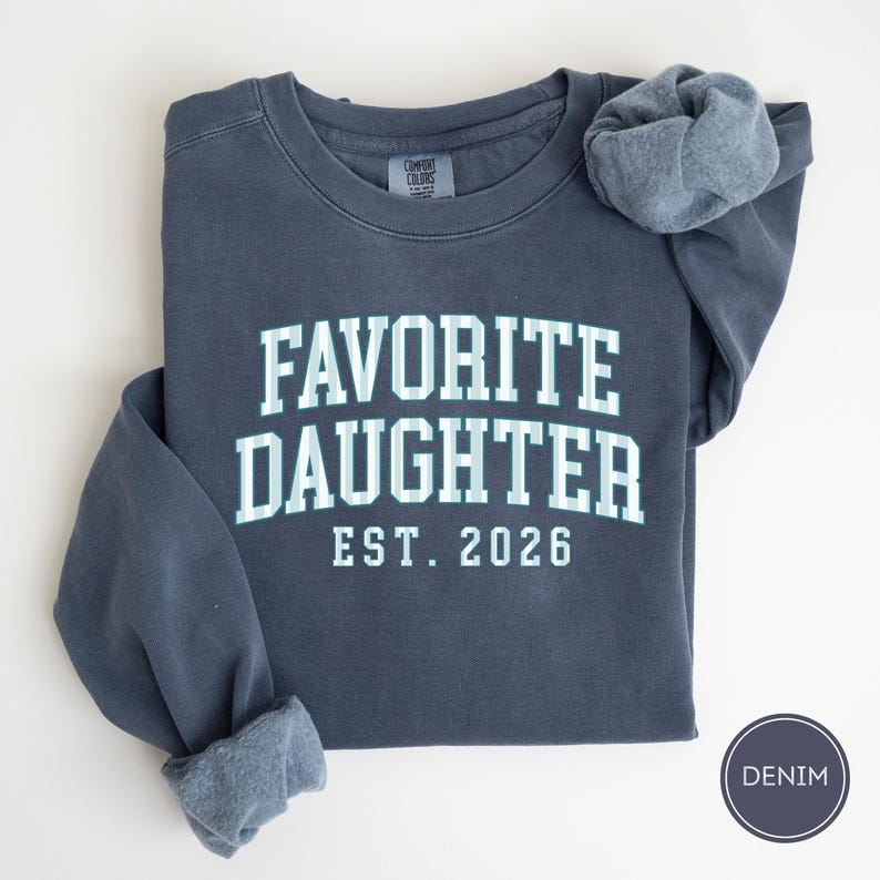 May include: A dark blue crewneck sweatshirt featuring the text FAVORITE DAUGHTER EST. 2026 in a light blue and white striped font. The garment has a Comfort Colors brand label at the inner neckline and a circular badge labeled DENIM.