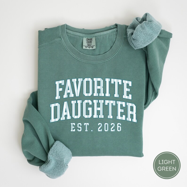 May include: A sage green crewneck sweatshirt featuring the white text FAVORITE DAUGHTER EST. 2026 in a collegiate font. The letters have a light blue and white checkered pattern. The garment has a ribbed collar and long sleeves with cuffs.
