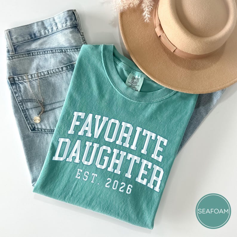 May include: A seafoam green short-sleeve t-shirt with white block lettering that reads FAVORITE DAUGHTER EST. 2026. The shirt is paired with light blue denim jeans, a tan wide-brimmed hat, and a delicate gold chain necklace with a small round pendant.