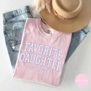 May include: A light pink t-shirt featuring the text FAVORITE DAUGHTER EST. 2026 in a purple and white striped collegiate font. The shirt is paired with light blue denim jeans, a tan wide-brimmed hat, and a delicate gold-toned necklace.