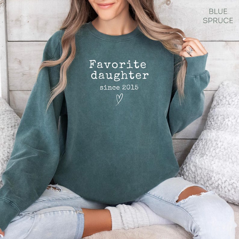 Pode incluir: Um moletom verde-azulado com o texto branco "Favorite daughter since 2015". O moletom tem gola redonda e mangas compridas. O texto est&aacute; centralizado no peito, com um pequeno cora&ccedil;&atilde;o abaixo.