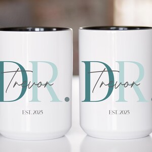 May include: Two white ceramic mugs with black handles and rims. Each mug features the text "DR Trevor" in teal and light blue, with "EST. 2025" below. The mugs are designed for beverages.