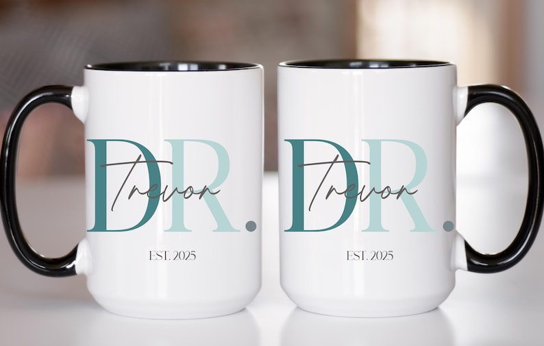 May include: Two white ceramic mugs with black handles and rims. Each mug features the text "DR Trevor" in teal and light blue, with "EST. 2025" below. The mugs are designed for beverages.