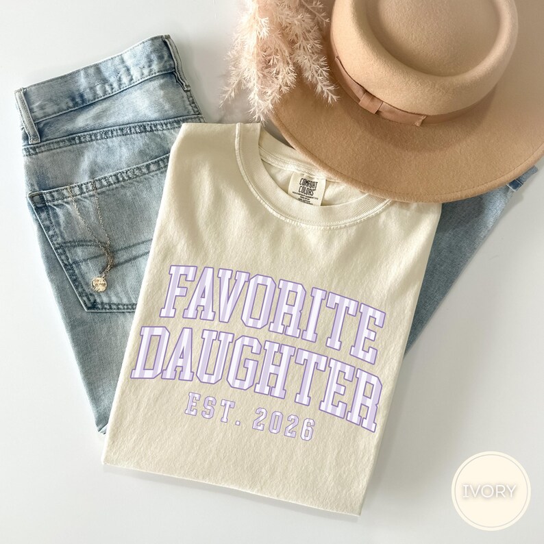 May include: An ivory t-shirt with the text FAVORITE DAUGHTER EST. 2026 in a light purple striped collegiate font, paired with light blue denim jeans, a tan wide-brimmed hat, and a delicate silver necklace with a small circular pendant.