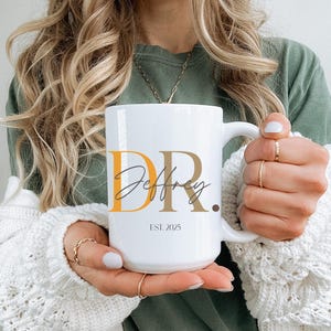 May include: White ceramic mug with the text "DR. Jeffrey" in a mix of orange, brown, and black fonts. The mug also has the text "EST. 2025". The mug is held by a person wearing a green shirt and a white sweater.