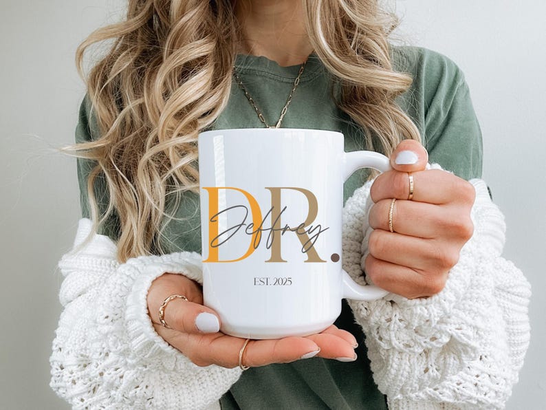 May include: White ceramic mug with the text "DR. Jeffrey" in a mix of orange, brown, and black fonts. The mug also has the text "EST. 2025". The mug is held by a person wearing a green shirt and a white sweater.
