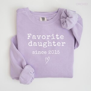 Pode incluir: Um moletom lil&aacute;s com a frase "Favorite daughter since 2015" em branco. Um pequeno cora&ccedil;&atilde;o &eacute; desenhado abaixo do texto. A pe&ccedil;a de roupa est&aacute; dobrada, mostrando as mangas e a gola.
