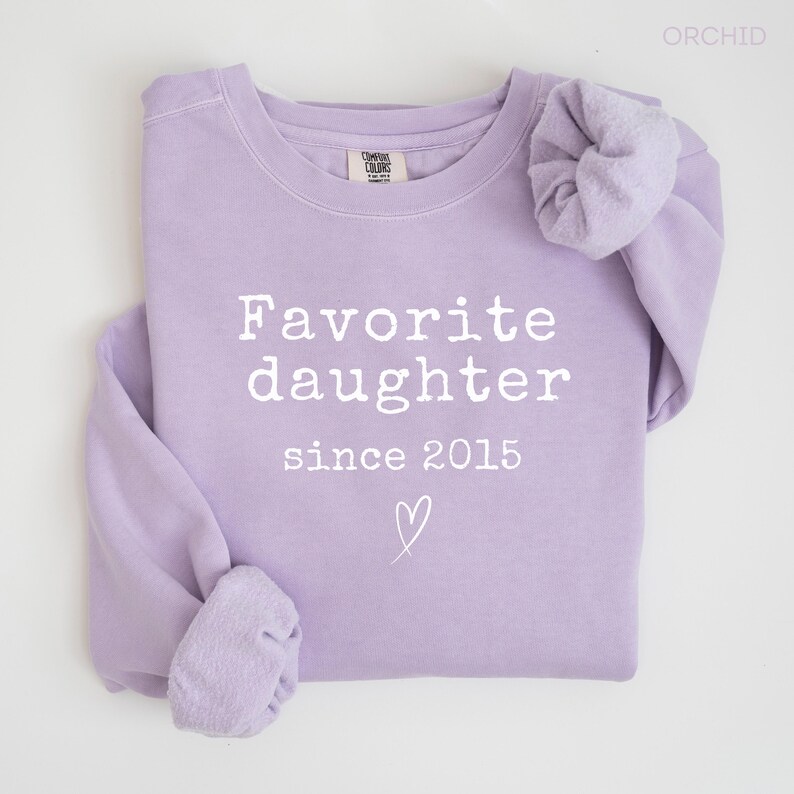 Pode incluir: Um moletom lil&aacute;s com a frase "Favorite daughter since 2015" em branco. Um pequeno cora&ccedil;&atilde;o &eacute; desenhado abaixo do texto. A pe&ccedil;a de roupa est&aacute; dobrada, mostrando as mangas e a gola.