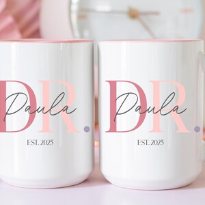 May include: Two white ceramic mugs with pink handles and interior rims. Each mug features the name "Paula" in a script font, with the letters "DR" in a stylized font. The text "EST. 2025" is printed below the name.
