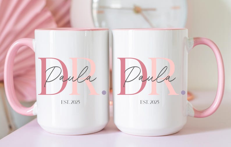 May include: Two white ceramic mugs with pink handles and interior rims. Each mug features the name "Paula" in a script font, with the letters "DR" in a stylized font. The text "EST. 2025" is printed below the name.