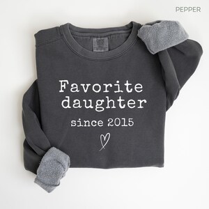 Pode incluir: Camisola cinza escuro com o texto branco "Favorite daughter since 2015" e um s&iacute;mbolo de cora&ccedil;&atilde;o. As mangas s&atilde;o dobradas. A camisola &eacute; feita de um material macio.